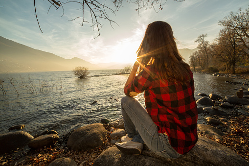 Young woman sits on a rock by the lake, watches the sunrise.