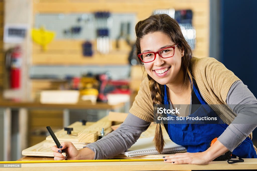 Artist working with wood in carpentry shop or makerspace Young adult Caucasian hipster woman is smiling and looking at the camera while working on a project. Artist is using wood to create something in a carpentry shop or makerspace. Woman is using a measuring tape and pencil to measure boards. She is wearing an apron and glasses. Accuracy Stock Photo