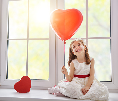 Sweet child girl with red heart. Little girl sitting on the window and holding balloon. Wedding, Valentine concept.