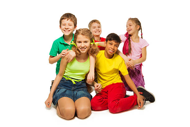 A small group of school aged children are seen sitting side-by-side outside.  They are each dressed casually and are smiling as they talk amongst each other.