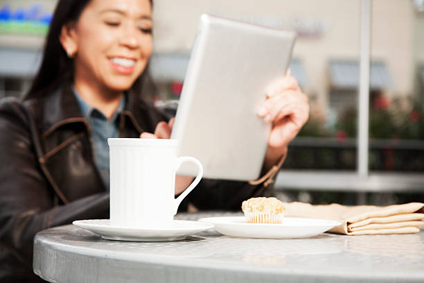Latin, Native American descent woman uses digital tablet at outdoor coffee shop or cafe. She scrolls through her social media apps while enjoying coffee and muffins.  Other shops behind the mid-adult woman. 