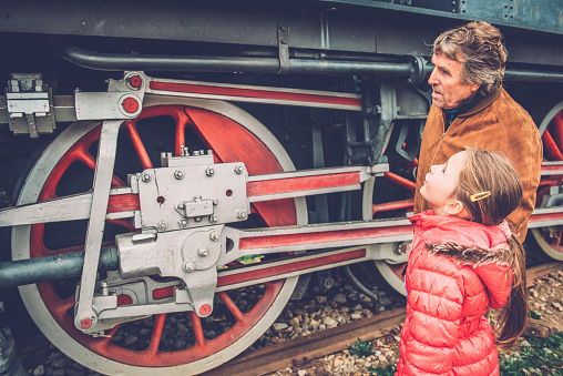 Girl with long brown hair in pink winter jacket and her grandfather admiring an old steam train at the Nova Gorica train station, Slovenia, Europe. They are looking at the wheel. Side view, close-up.  Nikon D800, full frame, XXXL.