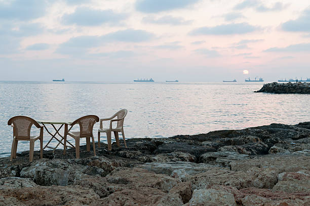 Three plastic chairs and a table, part of an outdoor cafe beside the Mediterranean Sea, wait customers at sunset in Tartus, Syria