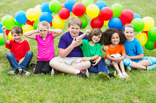 A multi-ethnic group of six children in the park, sitting on the grass in front of colorful balloons.  They are of mixed ages from 5 to 12 in summer camp.  The little girl in pink is making a face, but the rest are smiling.