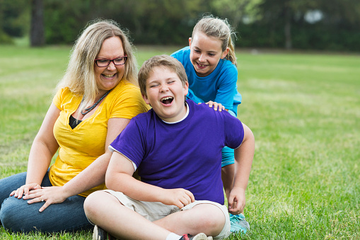 A mother sitting on the grass in the park, with her two children, a 12 year old son and his little sister.  The girl is standing behind her brother, making him laugh.