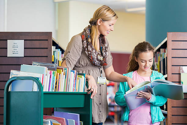 bibliotecário de escola primária, ajudando o aluno a ler um livro na biblioteca - librarian imagens e fotografias de stock