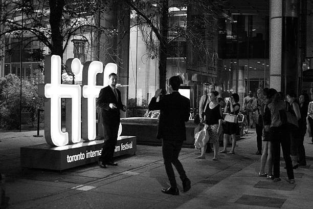 Tiff Toronto Сanada - September 16, 2015: Horizontal image of a man posing in-front of illuminated tiff sign, with male photo with cell phone and people on right side looking in. toronto international film festival stock pictures, royalty-free photos & images