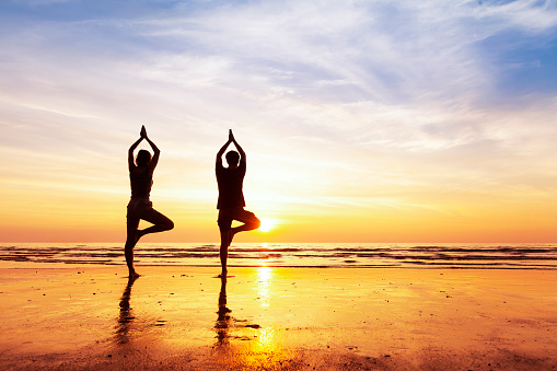 Two people practicing yoga tree position on the beach with beautiful sunset and reflection