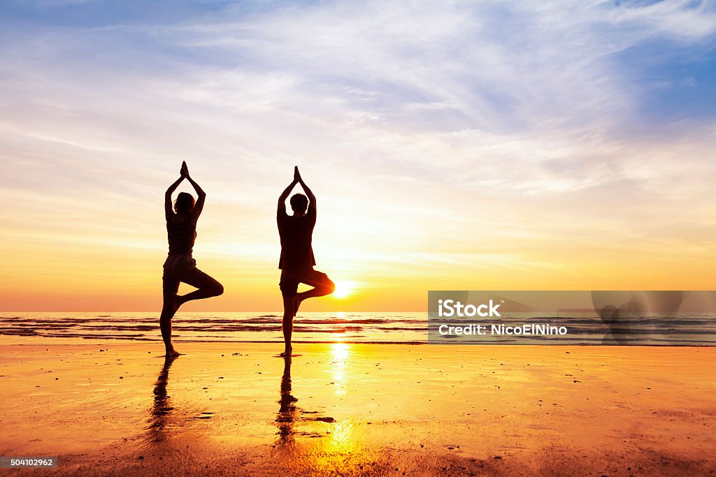 Two people practicing yoga tree position on the beach, sunset Two people practicing yoga tree position on the beach with beautiful sunset and reflection Yoga Stock Photo