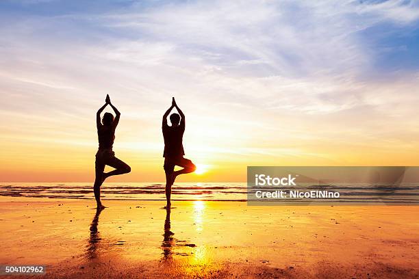 Two People Practicing Yoga Tree Position On The Beach Sunset Stock Photo - Download Image Now