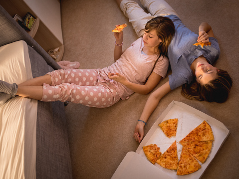 Two teen girls lying casually on a bedroom floor in their pyjamas with one resting on the other's stomach eating pizza from a take-away box