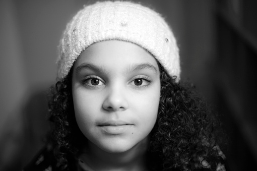 Stock photo of 9 years old girl who is wearing a bonnet with her curly hair, she is looking straight at the camera with a little smile. Post processed in black and white. Image taken indoors. This file has a signed model release.