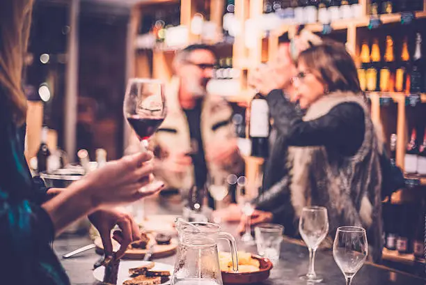 Woman holding a glass of red wine in the front, people talking and having fun in wine bar in the background, selective focus