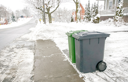 Two recycling bins and garbage awaiting pickup. A snow plow is at work in the background. Cover , snowy day. Positioning of bins appropriately located .
