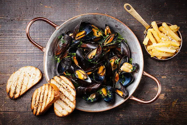 Mussels in copper cooking dish and french fries on dark wooden background