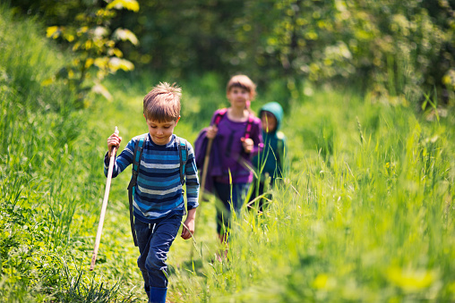 A sweet little girl pauses to pose for a photo during a family hike.  She is dressed casually, with a backpack on for her supplies and a walking stick in hand.  Her family can be seen standing in the background.