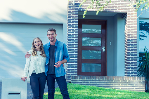 Couple standing in front of their new home. They are both wearing casual clothes and embracing. They are looking at the camera and smiling and he is holding a key to the house. The house is contemporary with a brick facade, driveway, balcony and a green lawn. The front door is also visible. Copy space