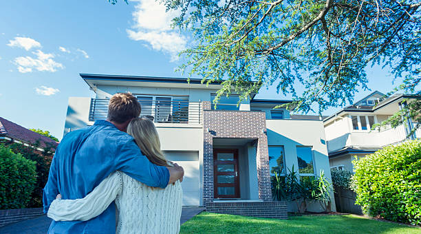 Couple standing in front of their new home. Couple standing in front of their new home. They are both wearing casual clothes and embracing. Rear view from behind them. The house is contemporary with a brick facade, driveway, balcony and a green lawn. The front door is also visible. Copy space buying a new house