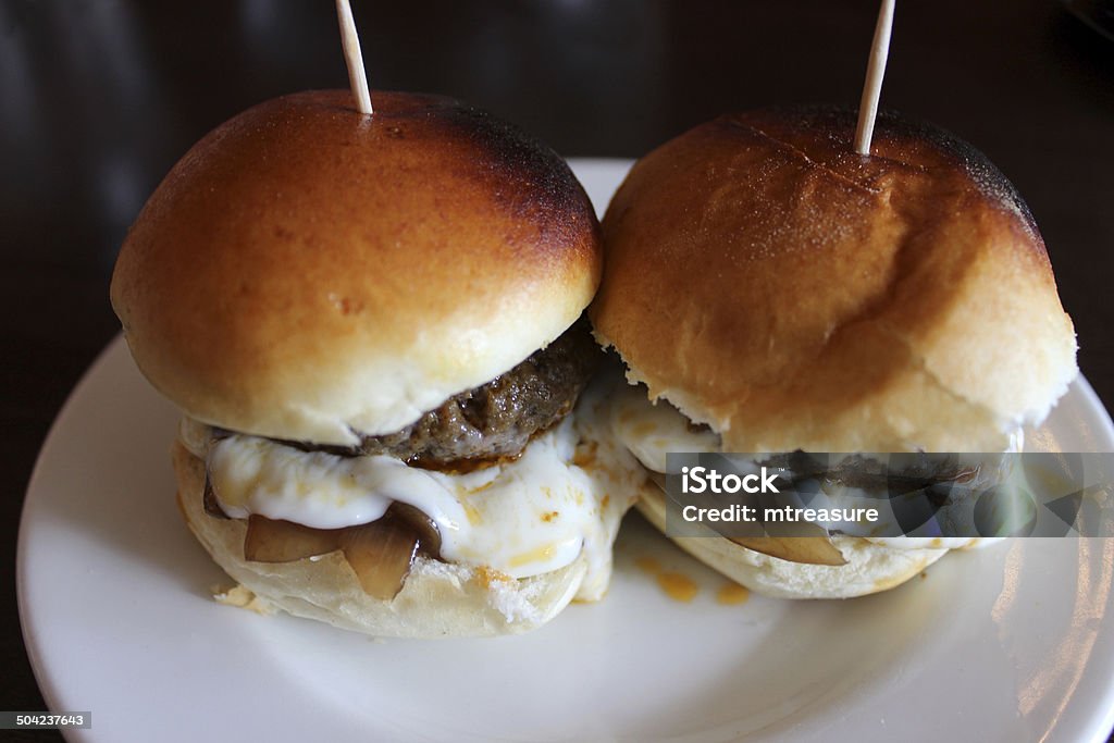 Image of mini beef burgers / mini burgers with cheese, tapas Photo showing a couple of mini beef burgers is small baps / bread rolls, served with a slice of cheese and mayonnaise, on a white plate.  The burgers were part of a meal at a Spanish tapas bar, together with other small dishes, and are pictured held together with wooden cocktail sticks. Appetizer Stock Photo