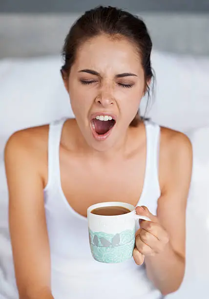 A young woman giving a big yawn while sitting with a cup of coffee in bed