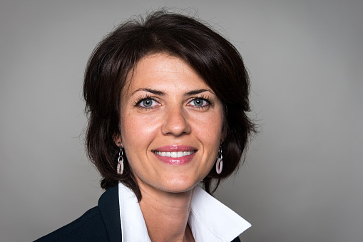 Smiling, Mature Businesswoman:A portrait of a smiling, mature businesswoman from the neck up. She has blue eyes and shoulder length brownish-black hair that is parted down the middle. She wears a white collared shirt and blue blazer, dangling earrings, and a soft pink lipstick. on gray background, studio shot. isolated on gray.