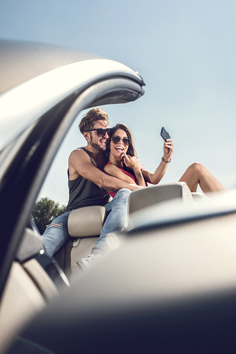 Young happy couple sitting on convertible car and having fun while taking a selfie with mobile phone.