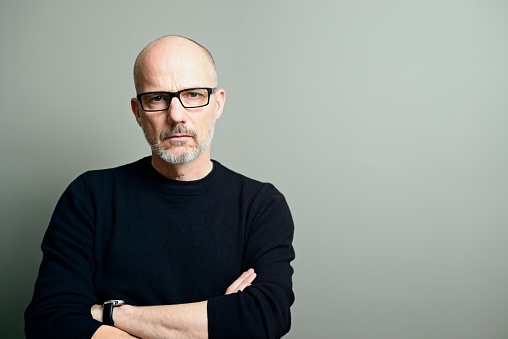 A handsome, balding mature man with arms crossed, wearing black-rimmed glasses, black t-shirt and sweater, looks seriously at the camera.