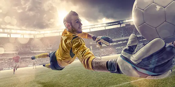 A close up mid action image of a professional soccer goalkeeper diving with arm out towards camera about to save football. The action occurs during a football game in a generic outdoor soccer stadium at sunset under a dramatic evening sky. The keeper is wearing generic unbranded soccer kit. 