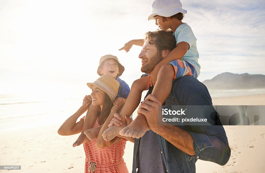 Look! I'm taller than you! A happy couple carrying their two young sons on their shoulders while they walk on the beach Adult Stock Photo