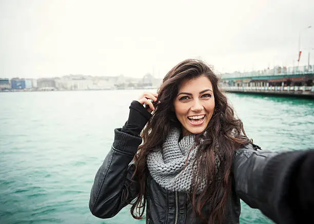 Lovely young girl with long curly dark hair is smiling brightly and looking at camera, as if making a selfie. The image is made with a wide angle lens. Bosphorus waters, a panoramic Istanbul city view and a bridge are in the background. Image contains plenty of copy space.