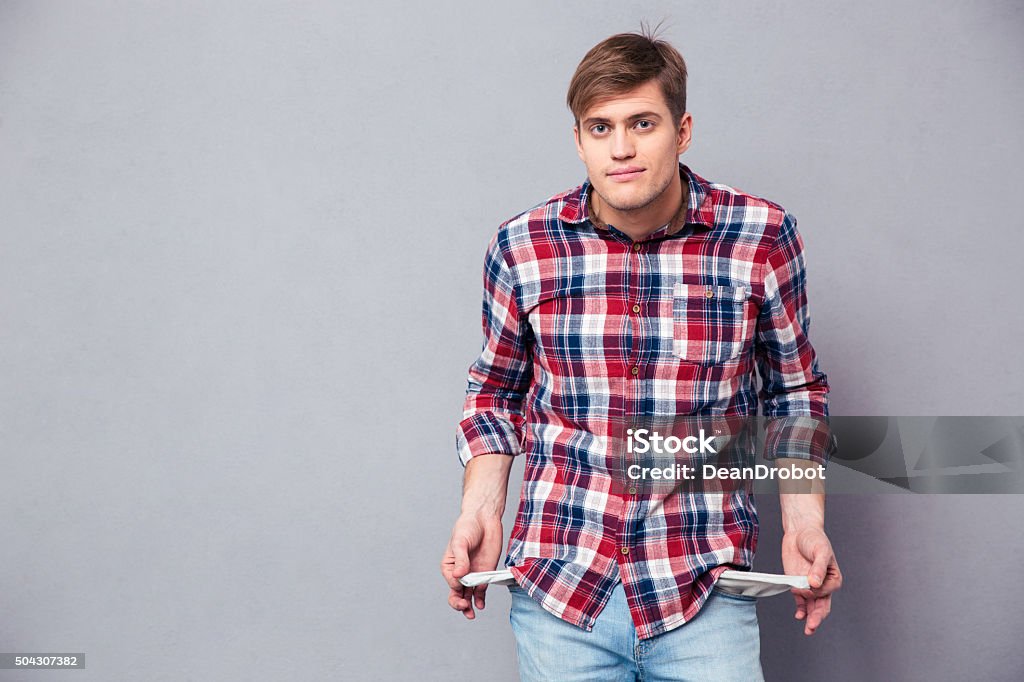 Poor handsome young man in checkered shirt showing empty pockets Poor handsome young man in checkered shirt and jeans showing empty pockets over grey background Pocket Stock Photo