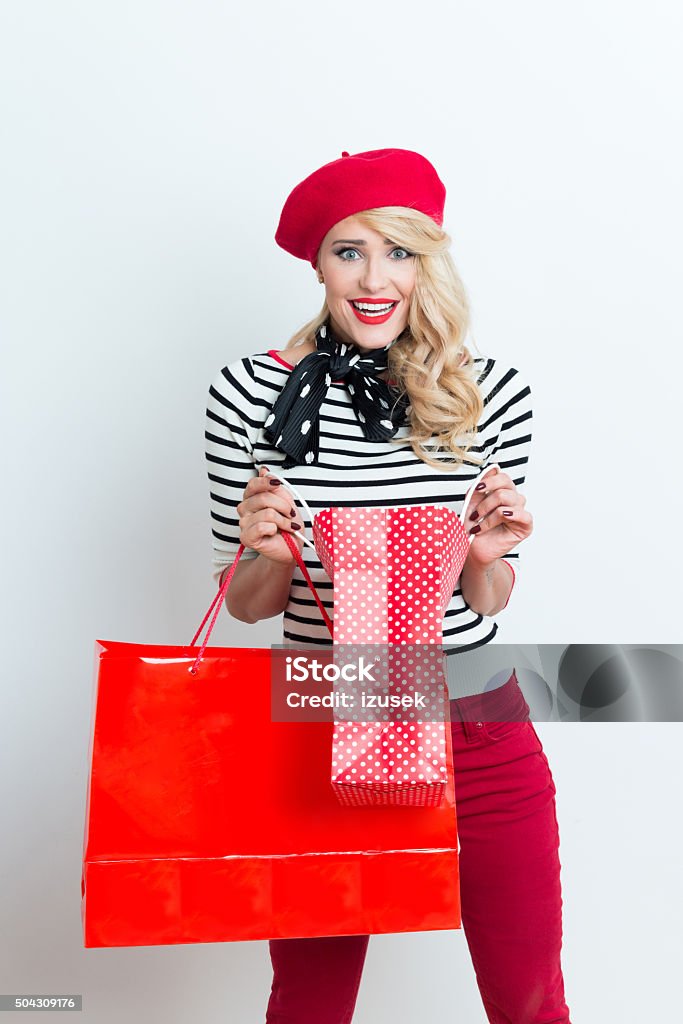 Surprised blonde french woman wearing red beret, holding shopping bags Portrait of surprised beautiful blonde woman in french outfit, wearing a red beret, striped blouse and neckerchief, holding shopping bags, staring at camera. Adult Stock Photo