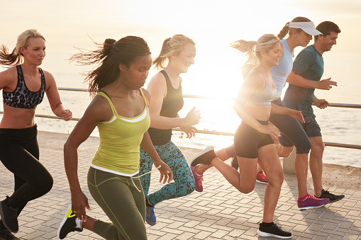 Portrait of healthy young men and women running race on seaside promenade. Group of young people sprinting outdoors at sunset.