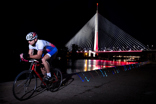 Man riding a bicycle along the river bank during evening hours. Bridge, river and city lights are in the background.