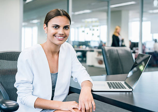Ethnic businesswoman at desk with laptop in modern office Young ethnic aboriginal Australian businesswoman seated at meeting room table by laptop computer modern office. Portrait looking to camera of a female business professional in Australia. aborigine woman stock pictures, royalty-free photos & images