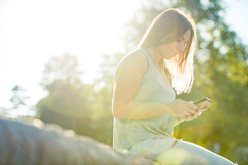 Portrait of attractive young woman sitting along the road using mobile phone on a bright sunny day. Sun is shining from back.