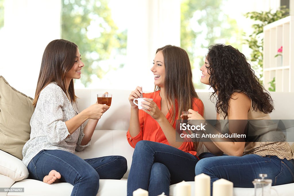 Three friends talking at home Three happy friends talking and drinking coffee and tea sitting on a couch at home Women Stock Photo