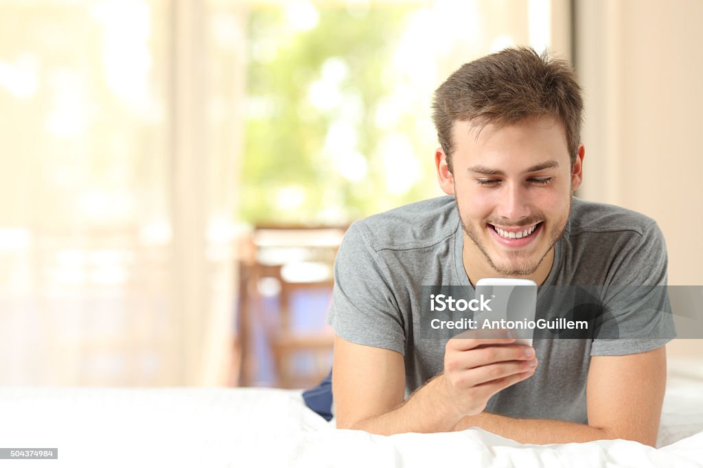 Guy using a mobile phone on a bed at home Front view of a guy using a mobile phone in the bedroom at home Adult Stock Photo