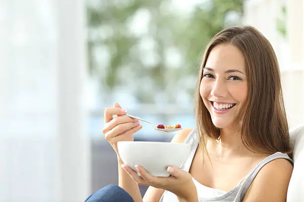 Casual happy woman dieting and eating cornflakes sitting on a couch at home