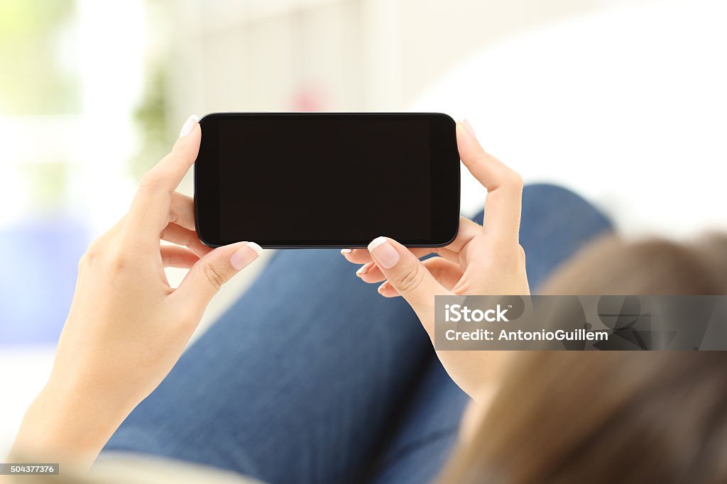 Woman watching media in a smart phone Back view of a close up of a woman hands watching media in a smart phone lying on a couch at home Smart Phone Stock Photo