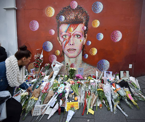 David bowie vigil London, UK - January 11th 2016: A woman leaves flowers at an impromptu shrine set up below a public mural of David Bowie in Brixton High Street, the district of South London where Bowie was born. David Bowie had died earlier on that day. brixton stock pictures, royalty-free photos & images