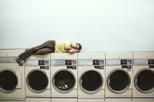 A man in 1980's style does his laundry at an old school laundromat.  He lays on top of the dryer machine and takes a nap while waiting for his clothes to dry.  Horizontal image with copy space.