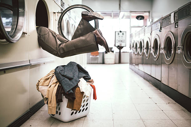 A man in 1980's style does his laundry at an old school laundromat.  He climbs into the dryer to reach some clothes in the very back, appearing to fall inside.  Horizontal image with copy space.