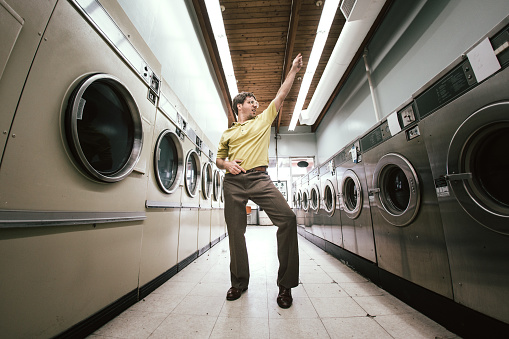 A man in 1980's style does his laundry at an old school laundromat, dancing disco style in the walkway between the washers and dryers while waiting for his clothing to finish drying.  Horizontal fisheye image with copy space.