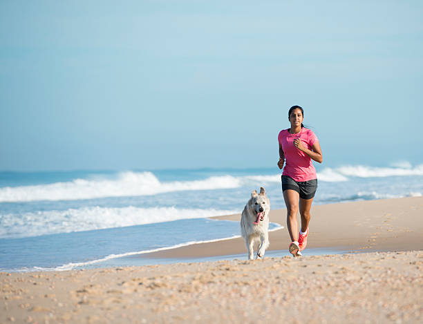 Young athletic woman running with her dog on sandy beach in the spring sunny day.