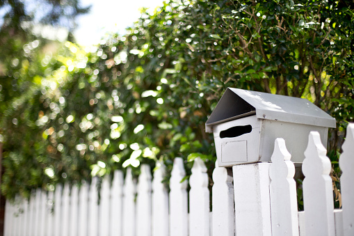 Mailbox attached to a white picket fence situated in front of a garden hedge.