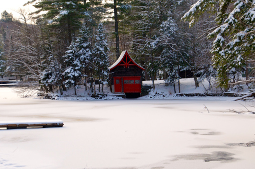 Red trimmed winter boathouse stands out on the shore of lake.