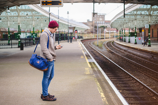 Young hipster styled man standing waiting for a train, using a smartphone. He is wearing a burgundy beanie hat and warm clothing.