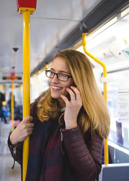 Young hipster styled woman standing on a train talking on a smartphone. She is wearing big framed glasses and is laughing.