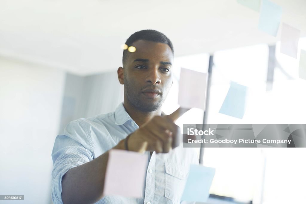 Creating a mind map of business strategies An African-American businessman looking at post-it notes stuck to a transparent wall African Ethnicity Stock Photo